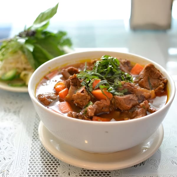 A bowl of Vietnamese beef stew with carrots, garnished with fresh herbs, and a side of greens.