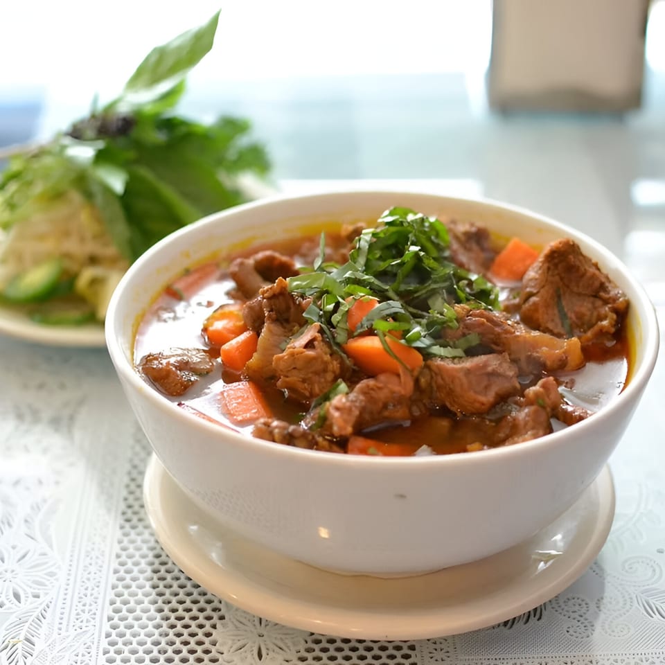 A bowl of Vietnamese beef stew with carrots, garnished with fresh herbs, and a side of greens.