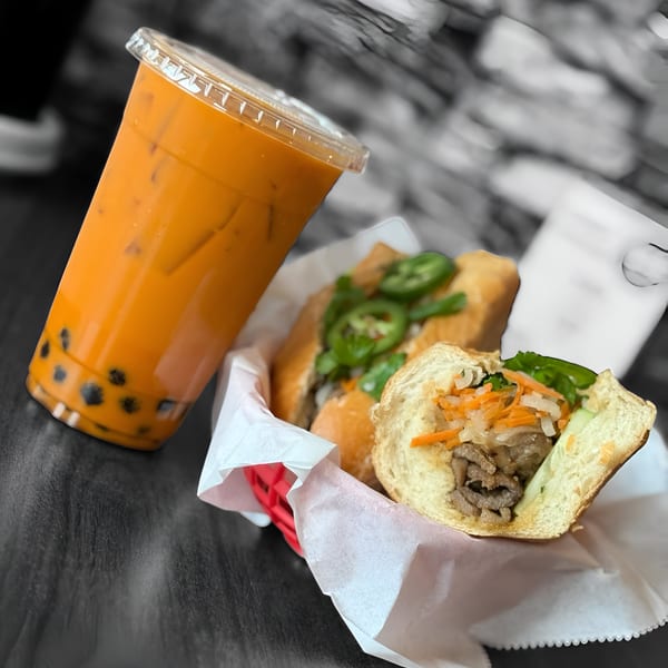 A Thai iced tea with boba next to two Vietnamese banh mi sandwiches in a basket.